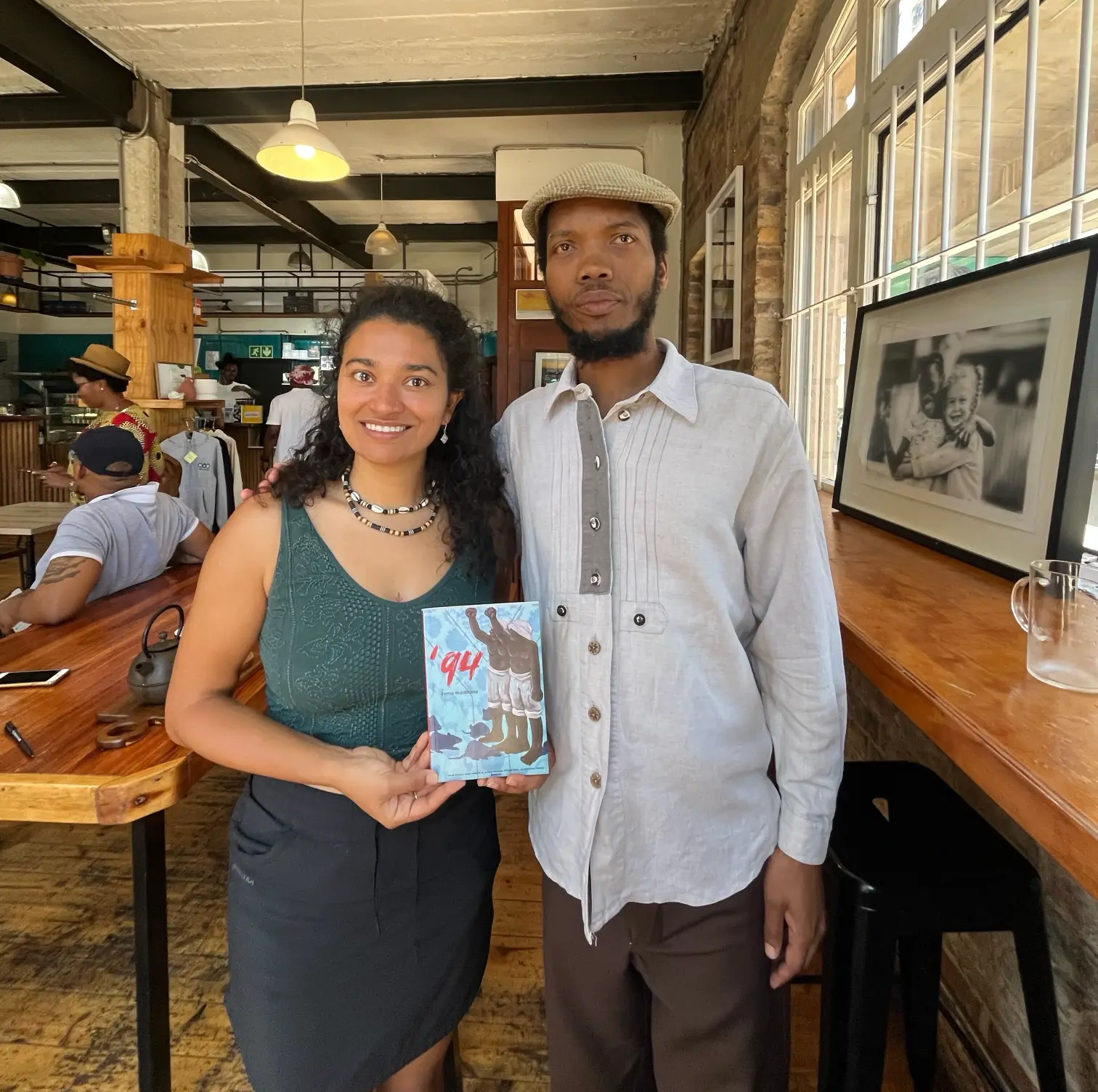Two people holding the poetry book titled '94 by Tshepo Madlingozi in a bright cafe in South Africa