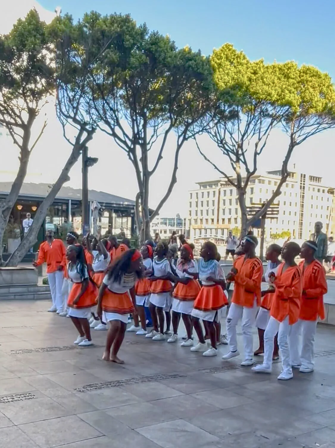 Youth choir and dance performers in orange and white uniforms performing outdoors in a South African plaza