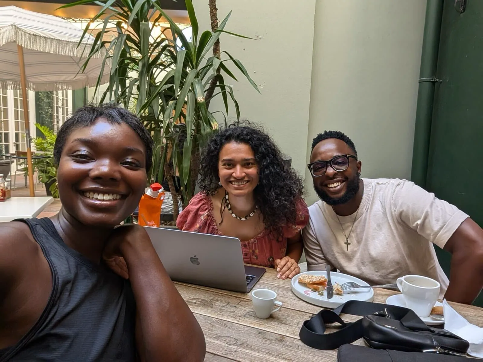 Three people smiling at an outdoor cafe table with a laptop, coffee, and food in South Africa