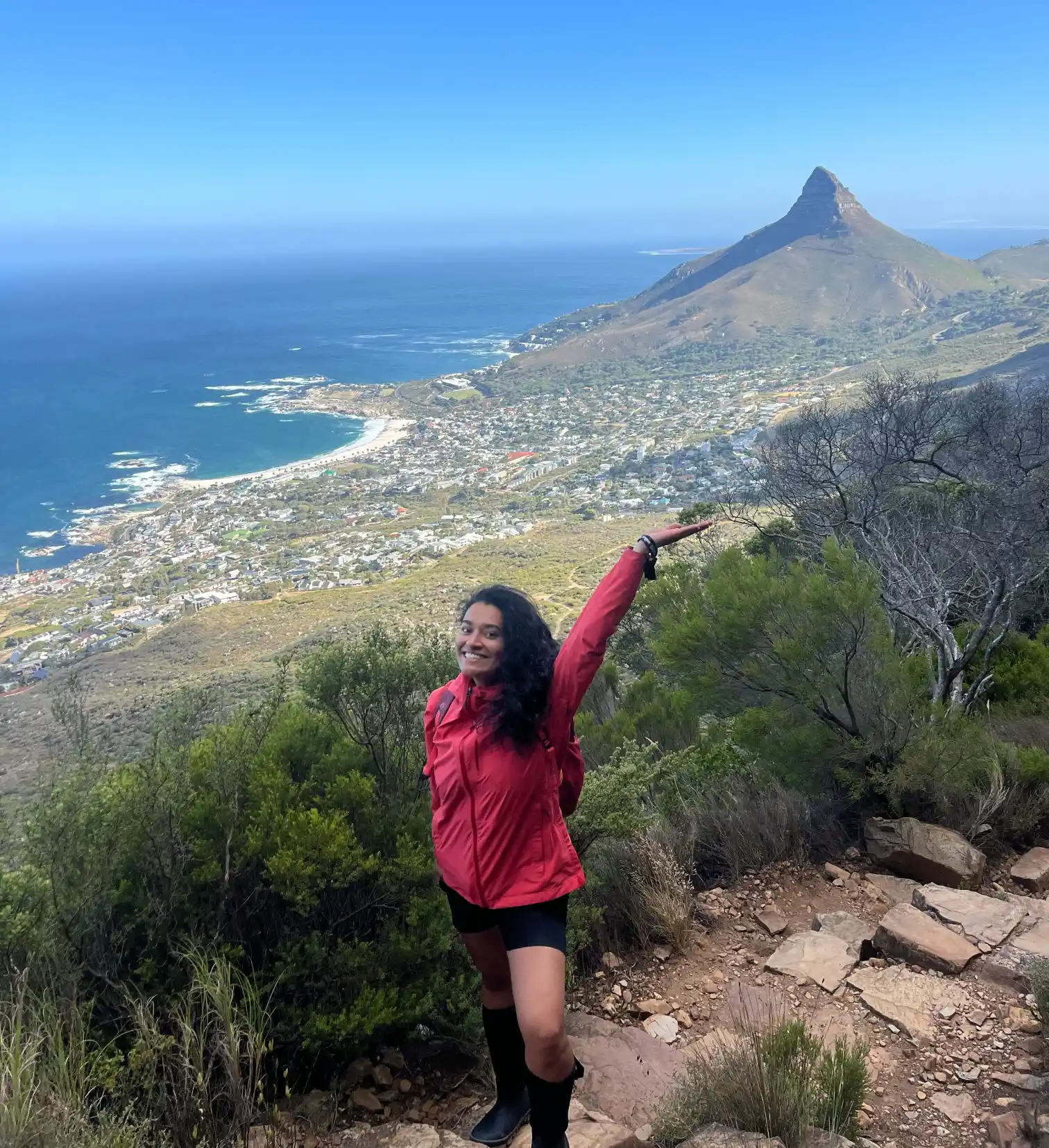 Hiker in a red jacket on a trail pointing toward Lion's Head with Cape Town and the coast in the background
