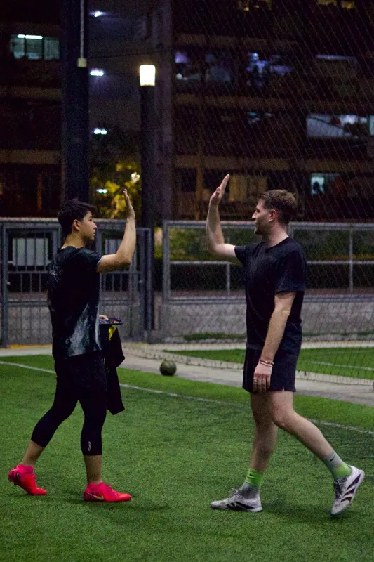 Two men share a high-five on a floodlit artificial turf football pitch at night in Bangkok
