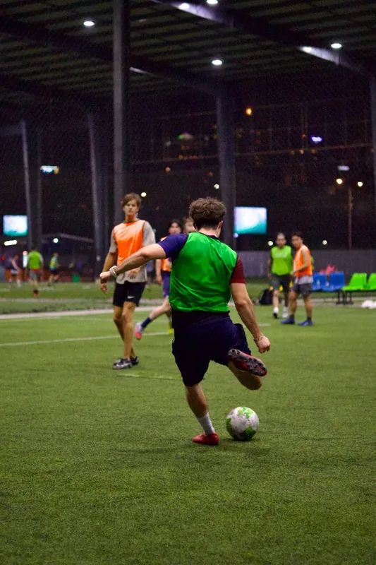 A player in a green bib prepares to kick a football during a night pickup game on artificial turf in Bangkok