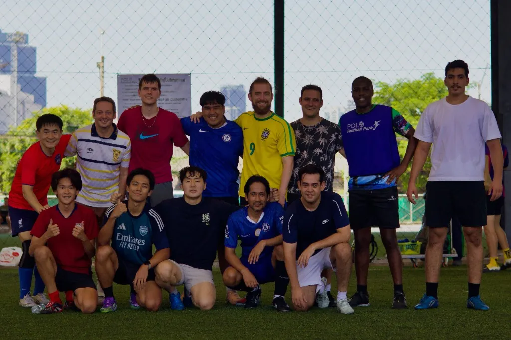 Thirteen men in mixed football jerseys pose for a team photo on an artificial turf pitch at Polo Football Park in Bangkok