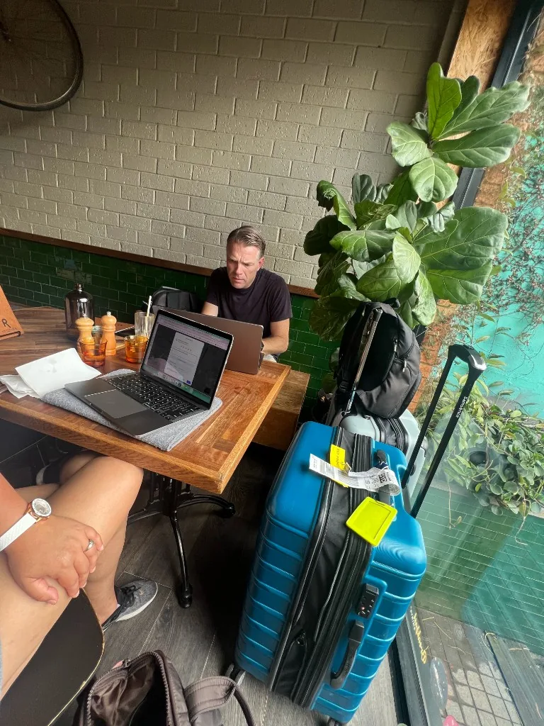 Nicole and her husband working on laptops at a cafe table surrounded by travel suitcases